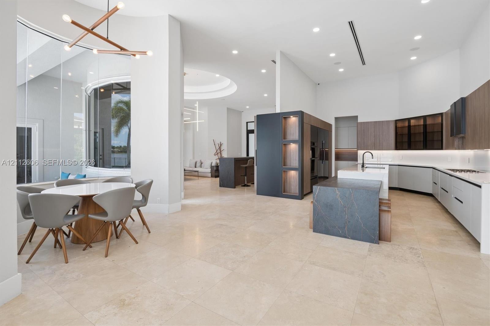 Open-concept interior featuring large-format stone tile flooring, high ceilings with recessed lighting, a modern kitchen with multi-toned cabinetry, an oversized stone-topped kitchen island, and a curved floor-to-ceiling window wall