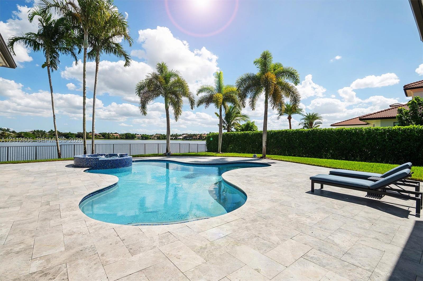 Freeform swimming pool with a raised spa, stone-paver decking, mature palm trees, and metal perimeter fencing overlooking a lake