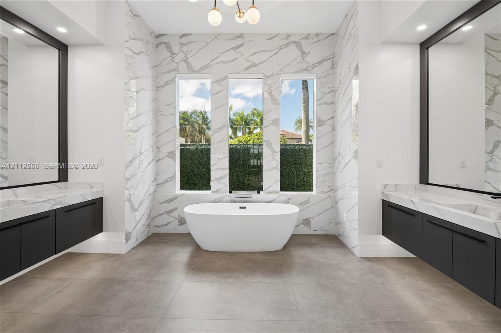 Freestanding soaking tub featuring large-format wall tiling, dual floating vanities with matte black hardware, framed wall-to-wall mirrors, and a modern multi-globe chandelier