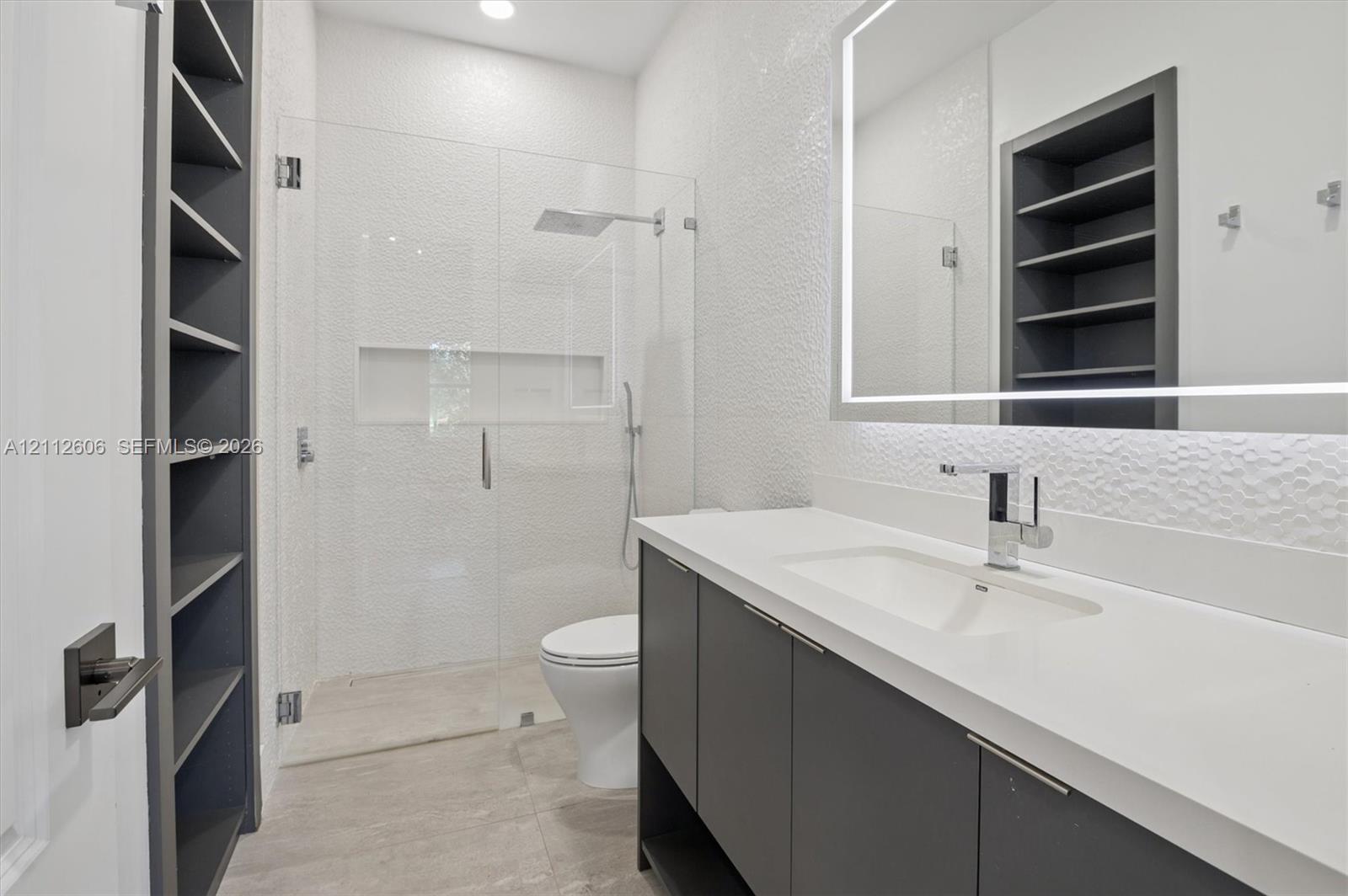 Floating vanity with solid-surface countertop and integrated sink, frameless glass-enclosed shower featuring built-in recessed niche, textured wall surfaces, illuminated vanity mirror, and integrated floor-to-ceiling shelving