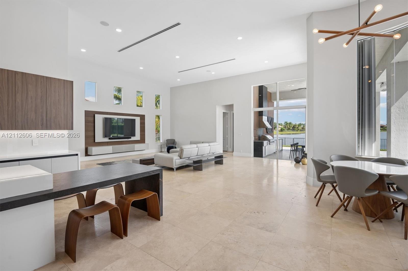 Open-concept interior featuring high ceilings, stone tile flooring, built-in media wall cabinetry, floor-to-ceiling glass paneling, and a contemporary multi-arm chandelier