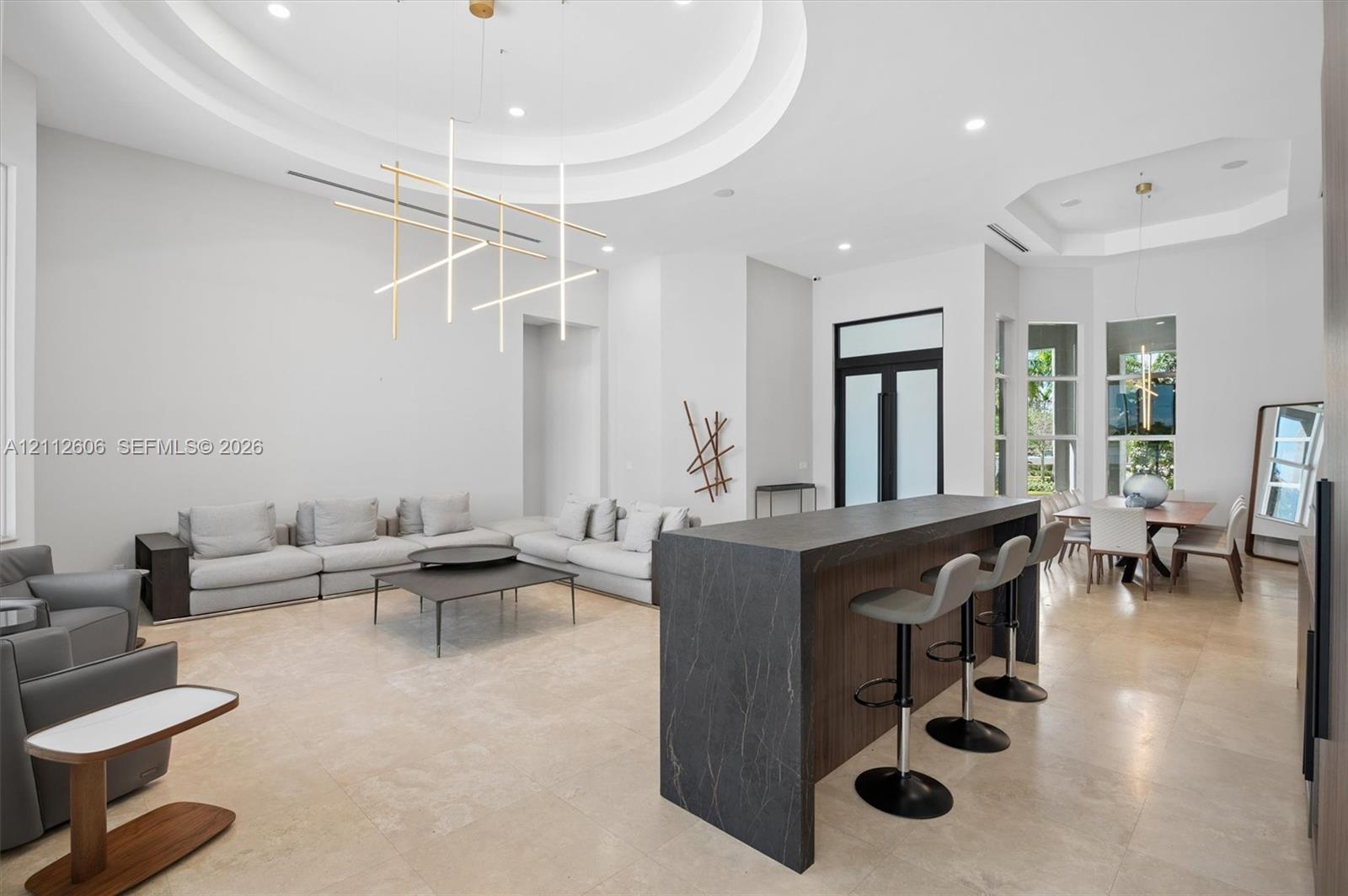 Open floor plan featuring high ceilings with recessed tray detailing, stone-finish flooring, a waterfall-edge kitchen island with dark cabinetry, and modern linear pendant lighting Open floor plan featuring high ceilings with recessed tray detailing, stone-finish flooring, a waterfall-edge kitchen island with dark cabinetry, and modern linear pendant lighting