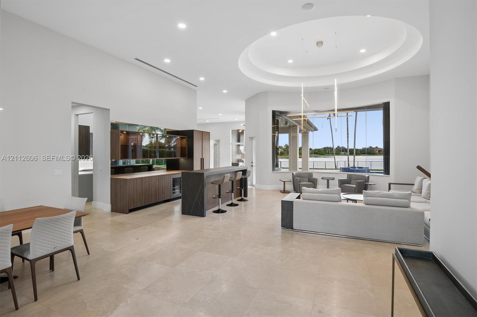 Open-concept interior featuring large-format tile flooring, a built-in wet bar with wood-finish cabinetry, an oval tray ceiling, recessed lighting, and expansive windows Open-concept interior featuring large-format tile flooring, a built-in wet bar with wood-finish cabinetry, an oval tray ceiling, recessed lighting, and expansive windows
