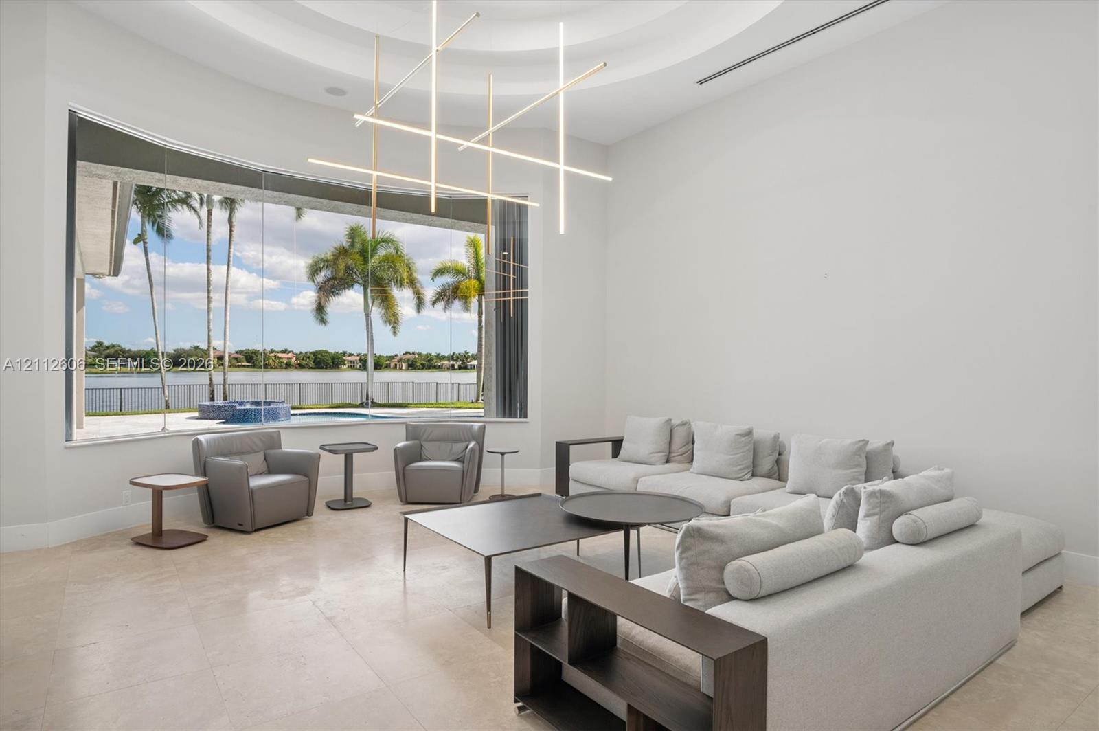 High-ceiling living area featuring large-format stone flooring, a circular tray ceiling, modern linear geometric chandelier, and floor-to-ceiling windows with waterfront scenery High-ceiling living area featuring large-format stone flooring, a circular tray ceiling, modern linear geometric chandelier, and floor-to-ceiling windows with waterfront scenery