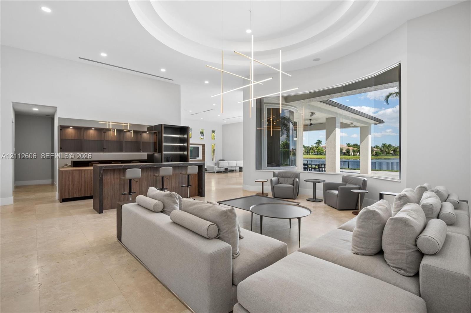 Open-concept space featuring a circular tray ceiling, large-format tile flooring, built-in wet bar with wood-finish cabinetry, contemporary linear chandelier, and expansive floor-to-ceiling windows Open-concept space featuring a circular tray ceiling, large-format tile flooring, built-in wet bar with wood-finish cabinetry, contemporary linear chandelier, and expansive floor-to-ceiling windows