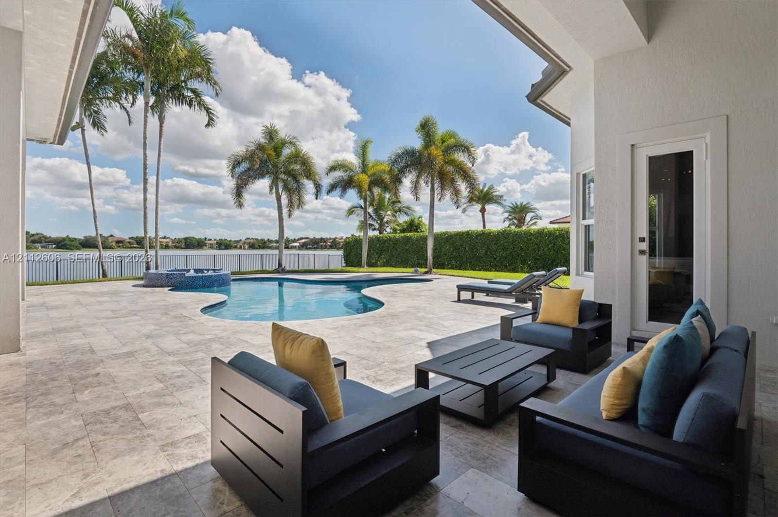 Covered patio featuring stone-tile flooring, free-form swimming pool with integrated spa, manicured perimeter hedge, and water backdrop