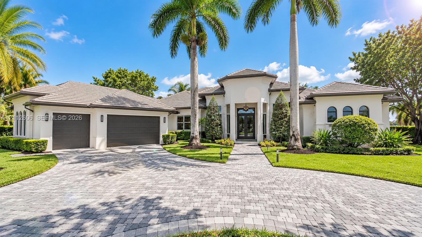 Stucco exterior with a multi-gabled tile roof, paver driveway, three-car garage, and formal entryway featuring double glass doors