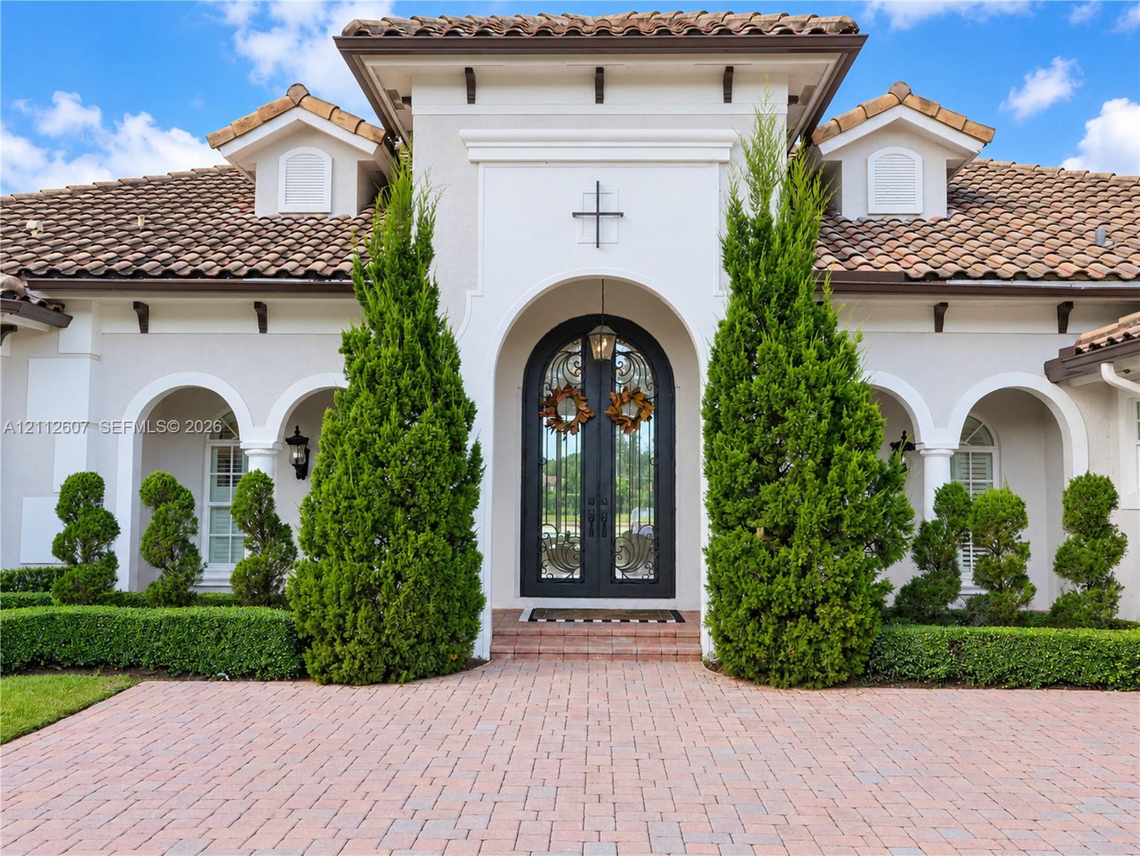 Stucco exterior featuring an arched entryway, double iron-detail glass doors, clay tile roof, and brick paver driveway