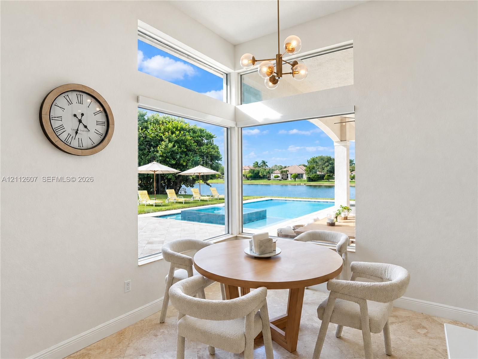 High-ceiling dining area featuring expansive corner windows, a multi-globe metal chandelier, and stone-finish flooring with outdoor water and pool access