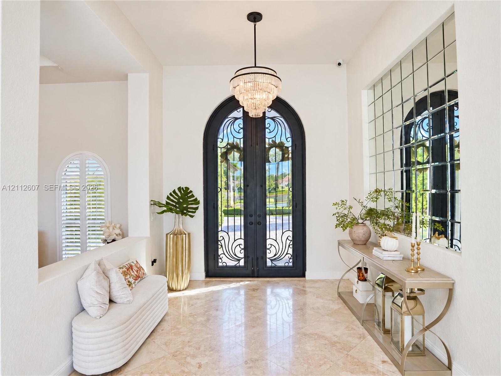 Foyer featuring arched double iron entry doors, light-toned stone flooring, a multi-tiered crystal chandelier, and an expansive grid-pattern wall mirror
