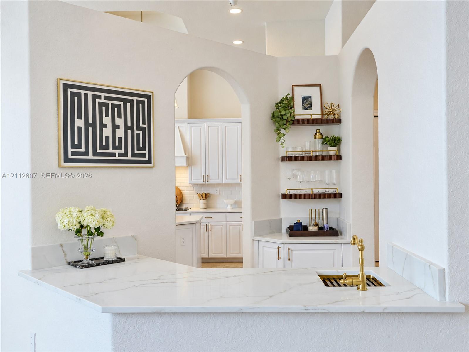 Built-in wet bar featuring stone-finish countertop, undermount sink with brass-finish faucet, and custom shelving, framed by architectural archways