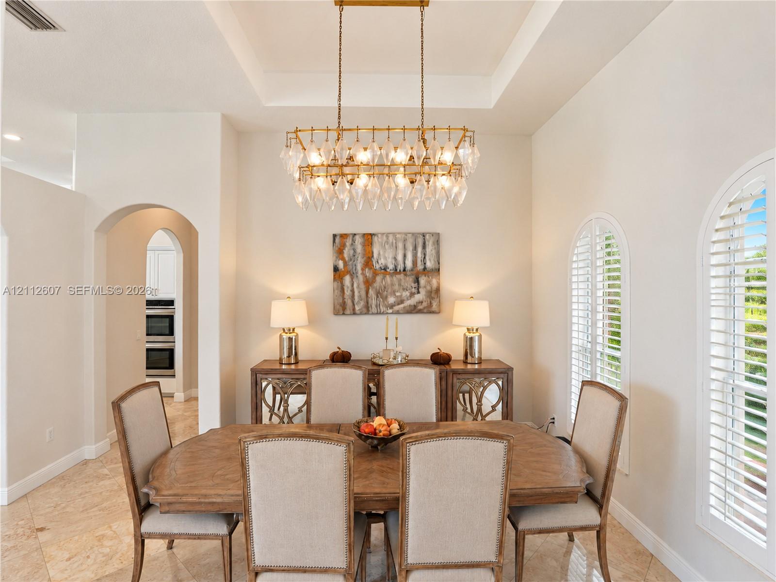 Tray ceiling with recessed lighting, tiered crystal chandelier, tile flooring, and arched windows featuring plantation shutters