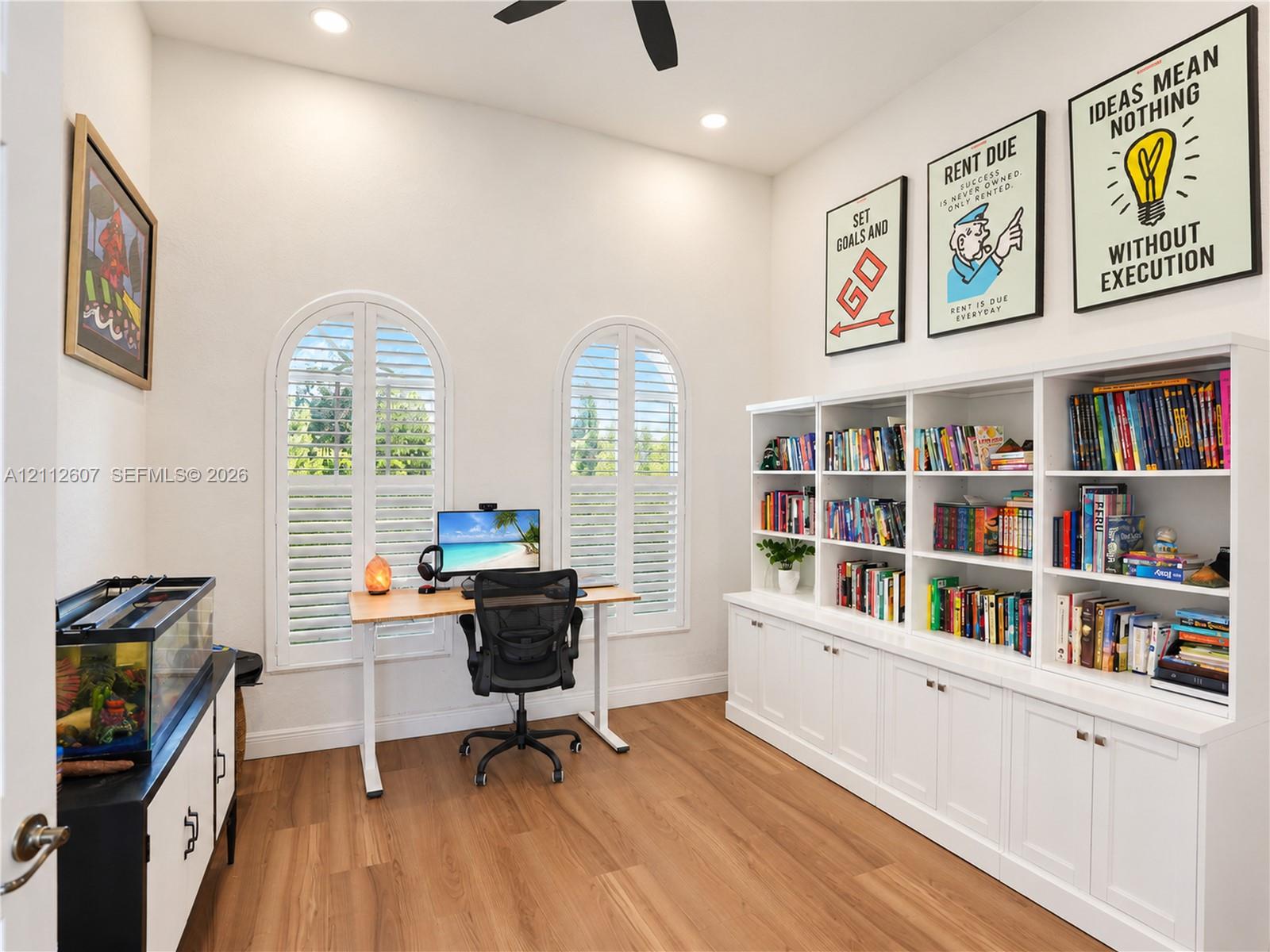 High-ceilinged room featuring wood-finish flooring, custom floor-to-ceiling built-in cabinetry, two arched windows with plantation shutters, and a ceiling fan