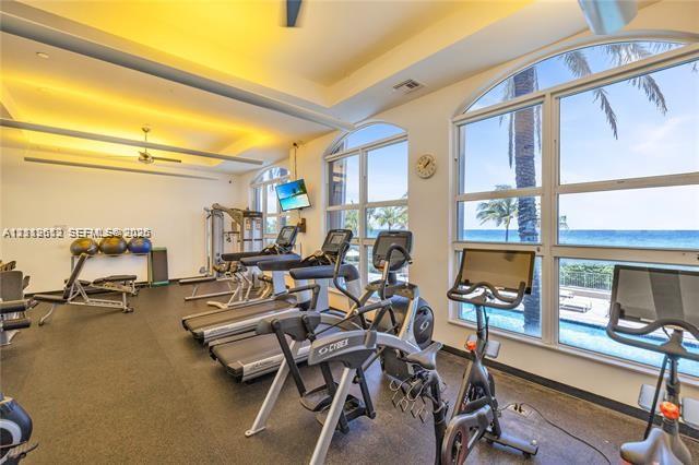 Fitness center featuring recessed tray ceiling lighting, floor-to-ceiling arched windows, and ocean backdrop