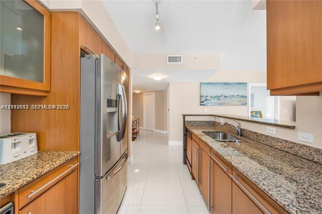 Kitchen featuring wood cabinetry, stone countertops, stainless steel refrigerator, tile flooring, and integrated sink