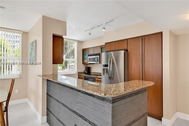 Kitchen featuring wood cabinetry, stone countertops, stainless steel appliances, a peninsula with textured paneling, and track lighting
