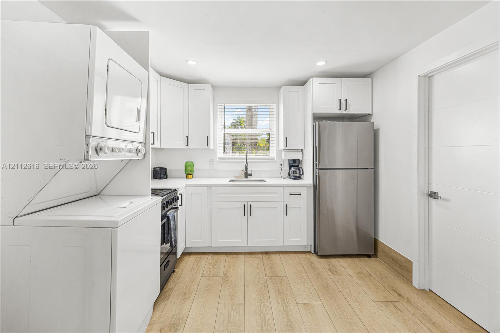 Kitchen featuring white shaker-style cabinetry, stainless steel refrigerator, solid-surface countertops, wood-finish flooring, and stacked laundry appliances