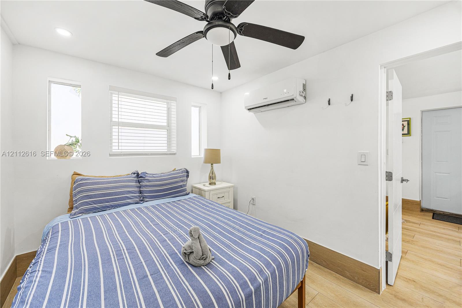 Room featuring wood-finish flooring, a wall-mounted ductless air conditioning unit, a ceiling fan with integrated lighting, and three windows equipped with horizontal blinds