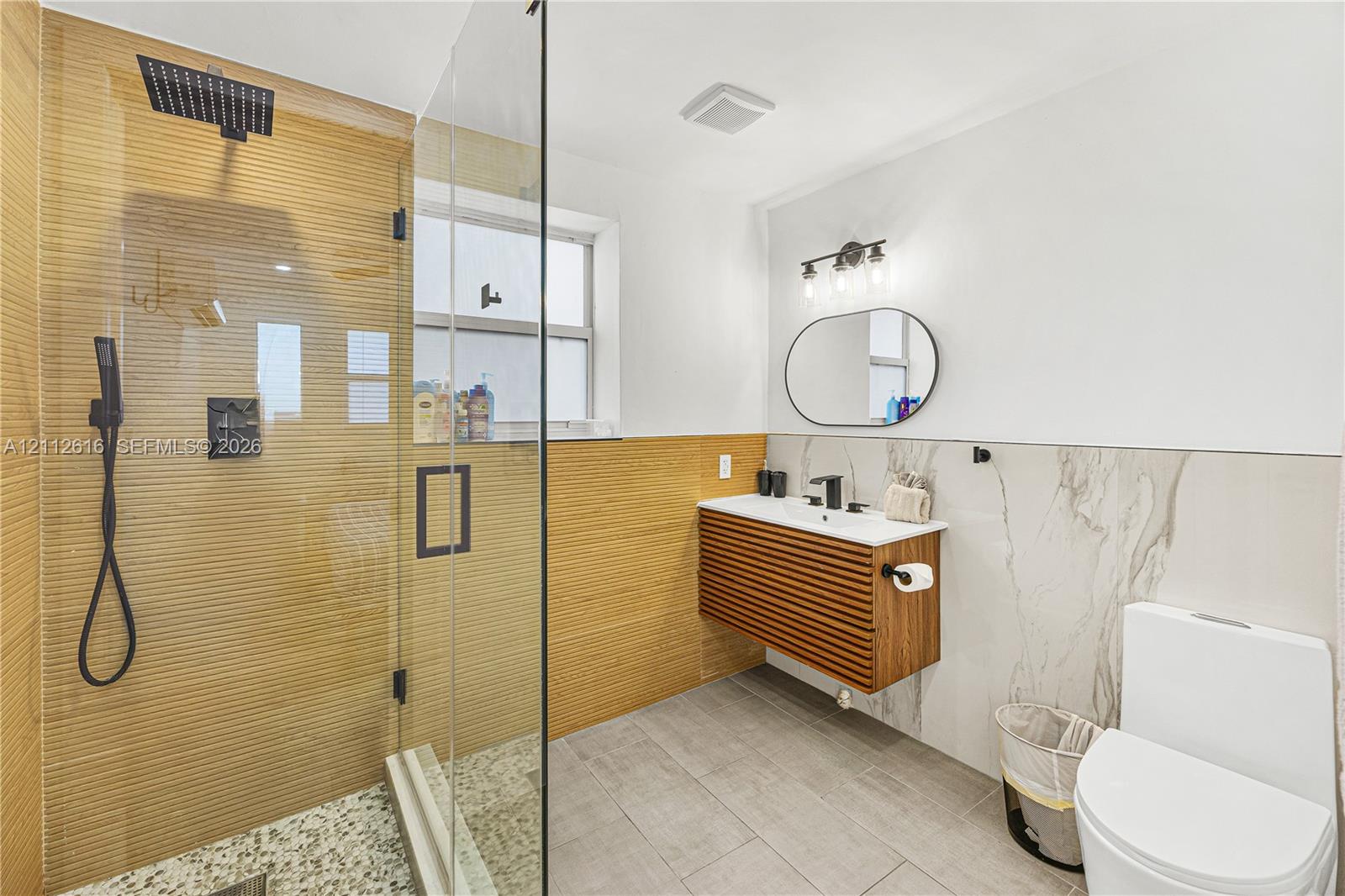 Modern bathroom featuring a glass-enclosed shower with a rainfall showerhead, textured wall tiling, a floating wood-slat vanity, a rectangular sink with matte black hardware, and large-format floor tiling
