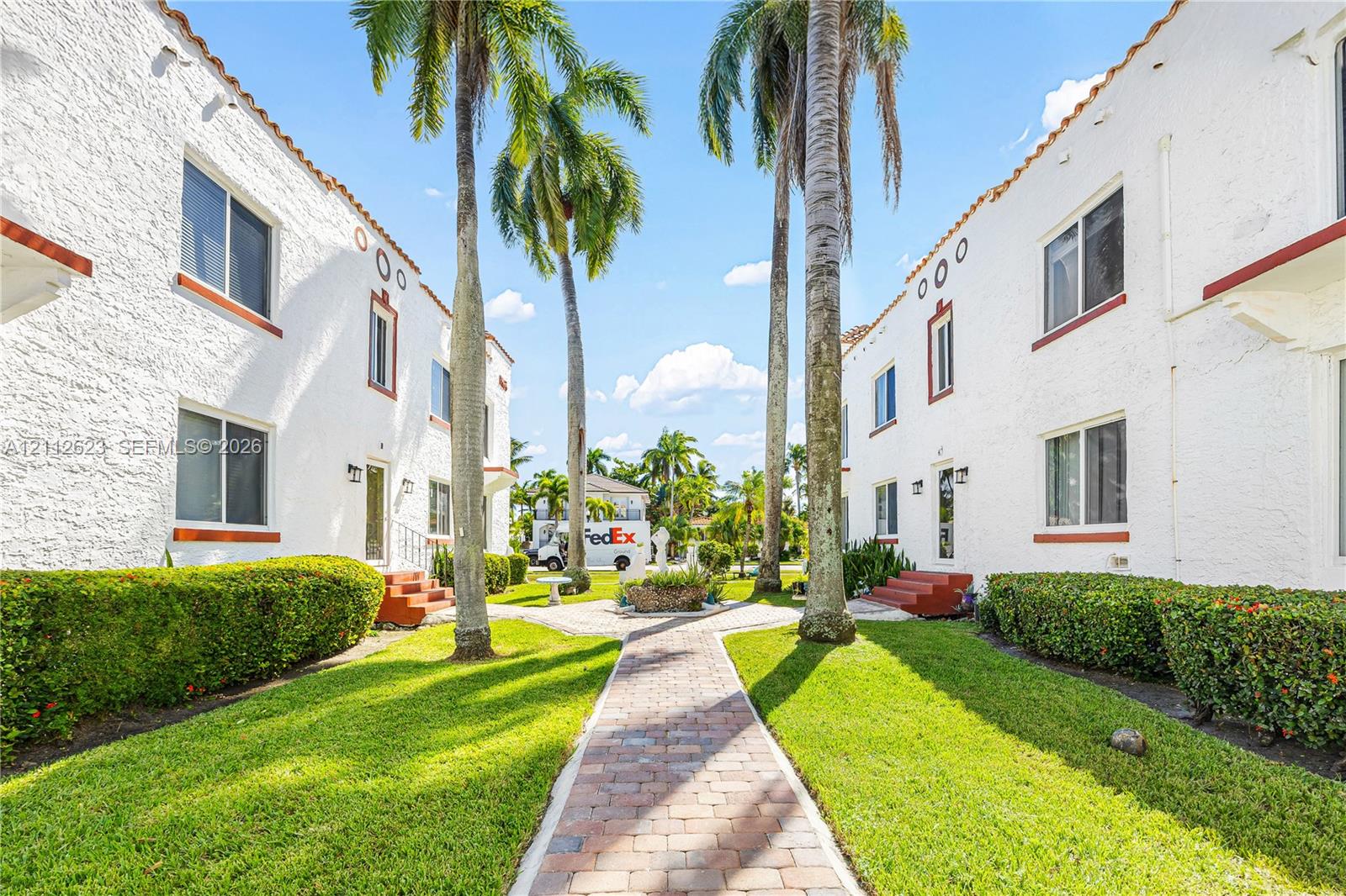 Stucco exterior featuring dual buildings with terracotta roof accents, manicured lawns, mature palm trees, and a central brick-paver walkway