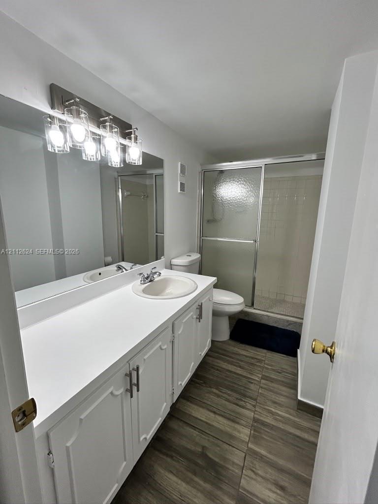 Bathroom featuring a wood-finish tile floor, extended white vanity with a solid-surface countertop, full-width mirror, multi-bulb wall-mounted lighting, and a glass-enclosed shower