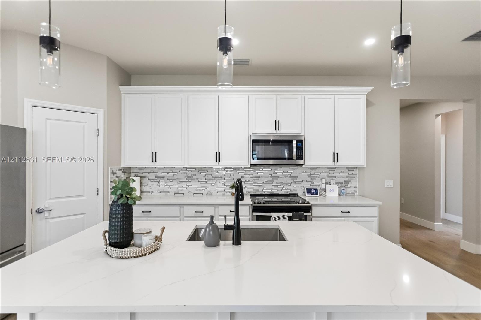 Kitchen featuring an oversized island with a solid surface countertop, white shaker-style cabinetry, a stone-patterned tile backsplash, matte black plumbing fixtures, and three cylindrical pendant lights