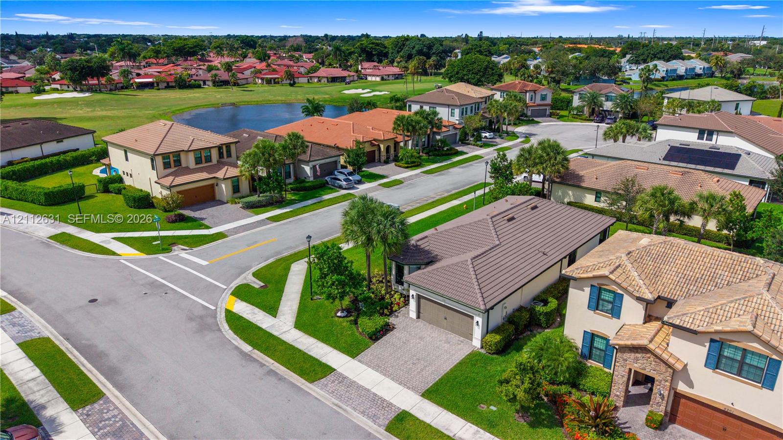 Aerial perspective of a residential neighborhood featuring paved sidewalks, street lighting, tile roof architecture, paved driveways, and proximity to an expansive grass common area with a water feature
