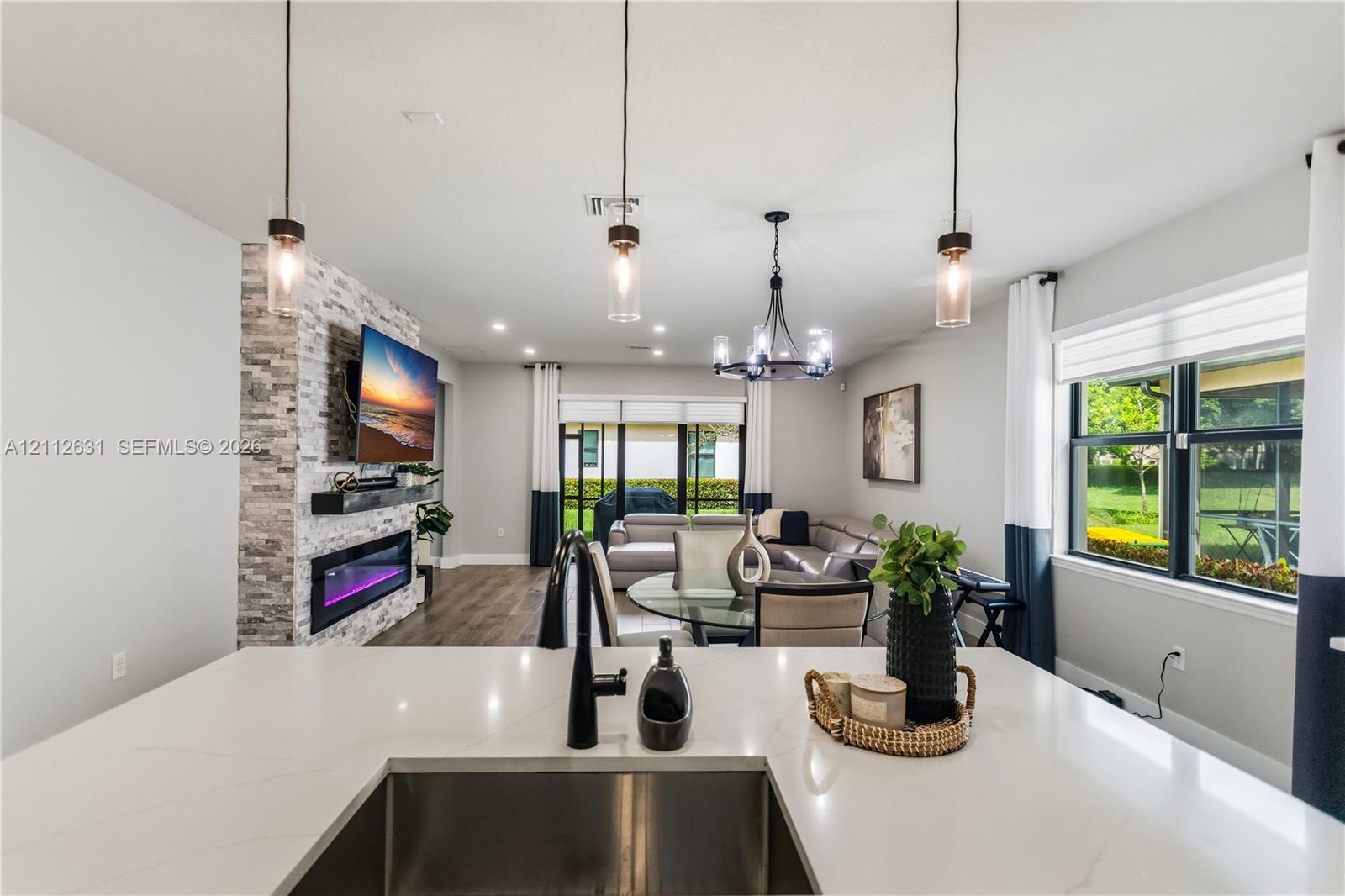 Kitchen island featuring a white stone countertop, undermount stainless steel sink, and matte black faucet, complemented by a floor-to-ceiling stone-veneer fireplace, wood-finish flooring, and multi-light pendant chandeliers