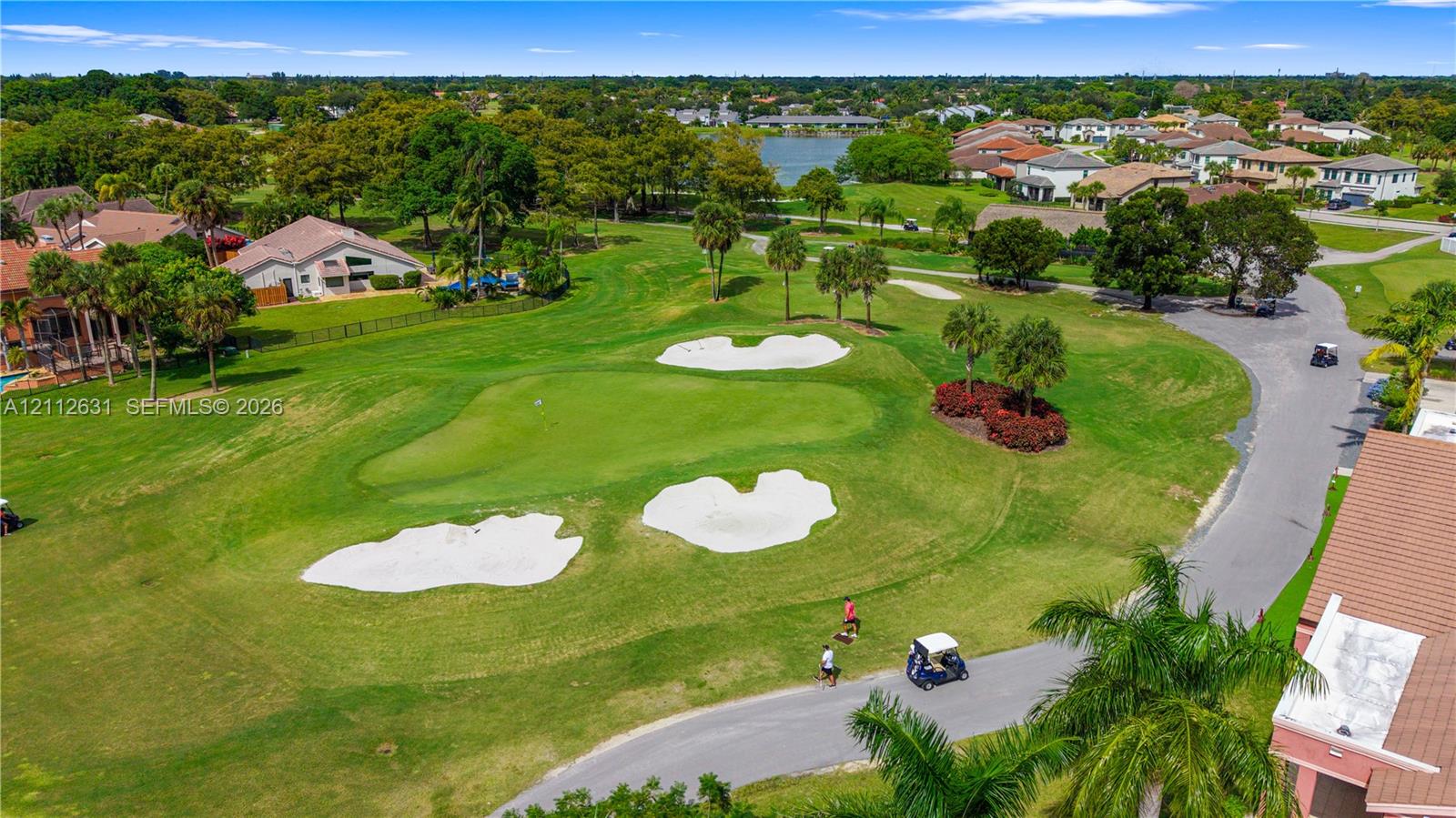 Aerial perspective of a golf course featuring manicured putting greens, sand bunkers, mature palm trees, and water frontage