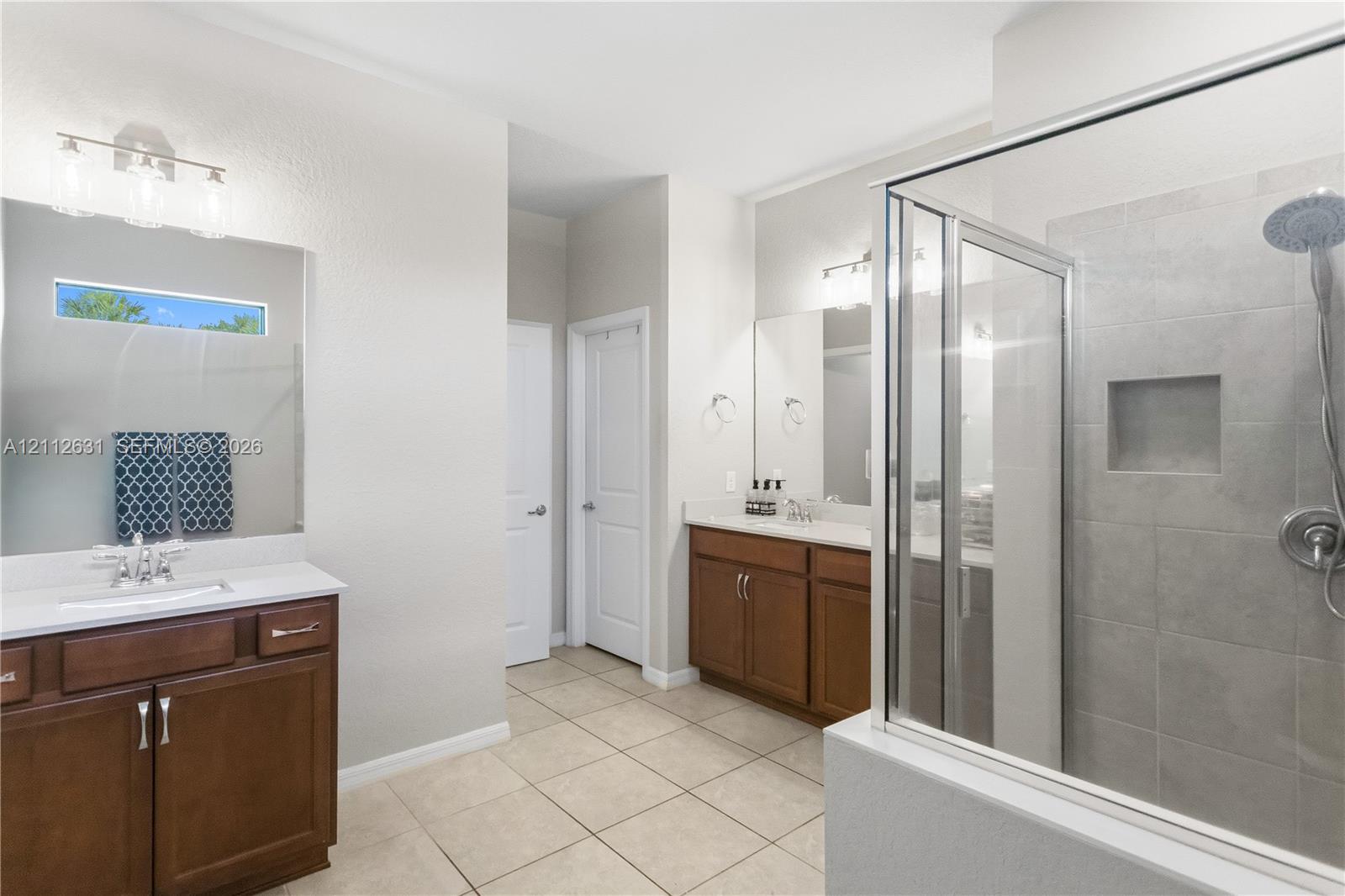 Bathroom featuring dual wood cabinetry vanities with light countertops, large wall-mounted mirrors, tile flooring, and a walk-in shower with tiled surround and built-in recessed niche
