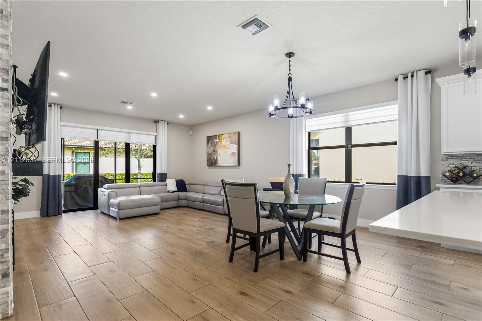 Open-concept living space featuring wood-finish flooring, recessed lighting, a multi-arm chandelier, large windows with horizontal blinds, and glass sliding doors
