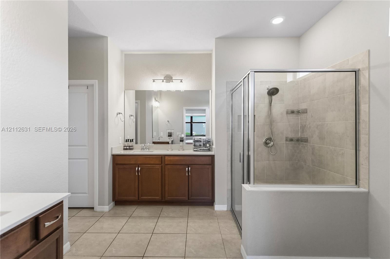 Dual-sink vanity with stone countertop, large wall-mounted mirror, glass-enclosed shower with tile surround, and tile flooring