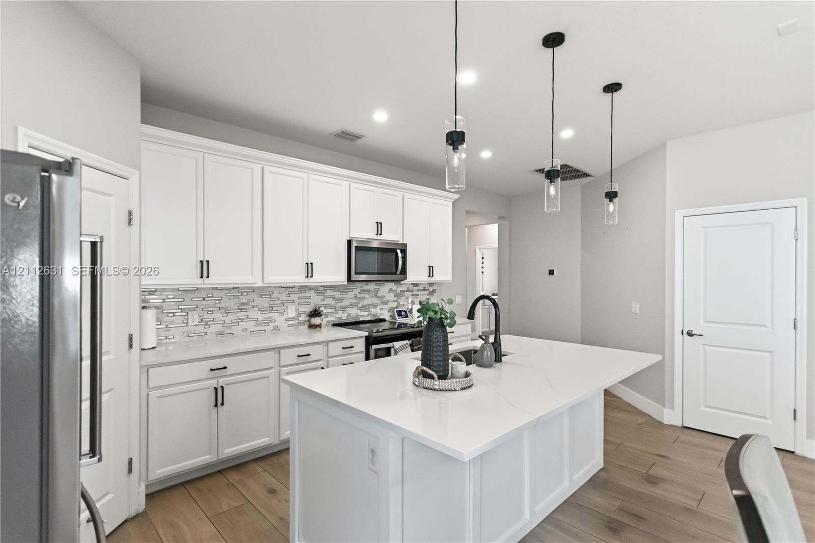 Kitchen featuring white shaker-style cabinetry, a large central island with a solid-surface countertop, linear tile backsplash, and three glass pendant lights