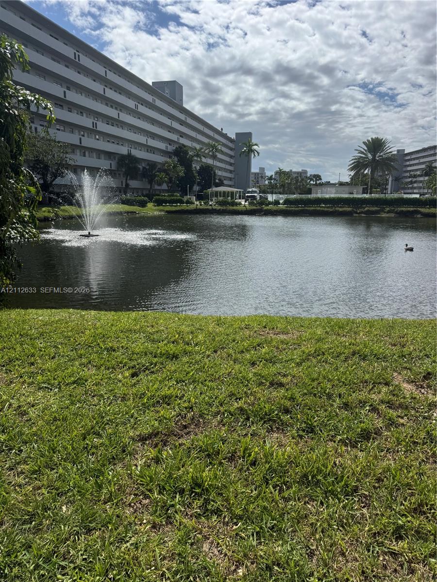 Manicured lawn encompassing a central pond with a mechanical fountain, framed by palm trees and multi-story structural exteriors