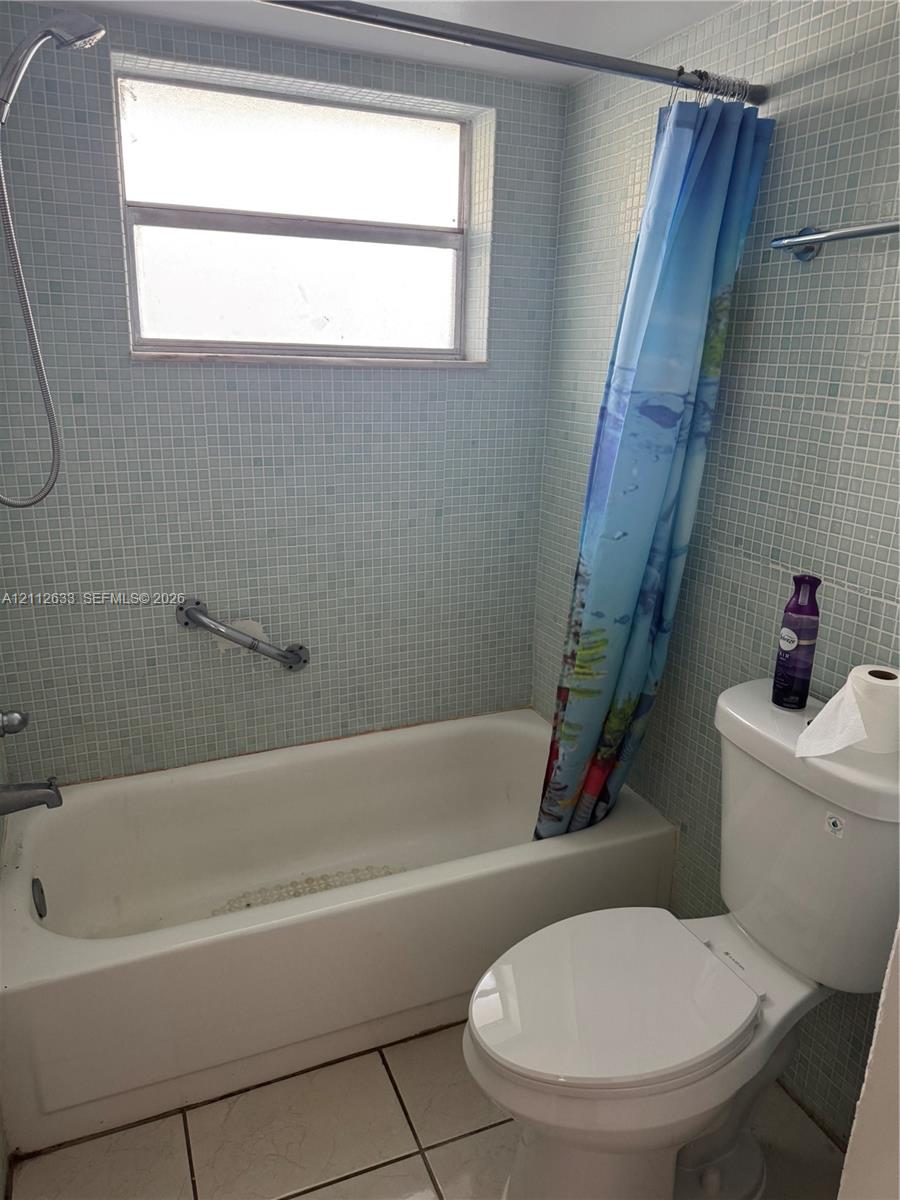 Full bathroom featuring light blue mosaic wall tile, white soaking tub with integrated shower head, horizontal window, and white floor tile