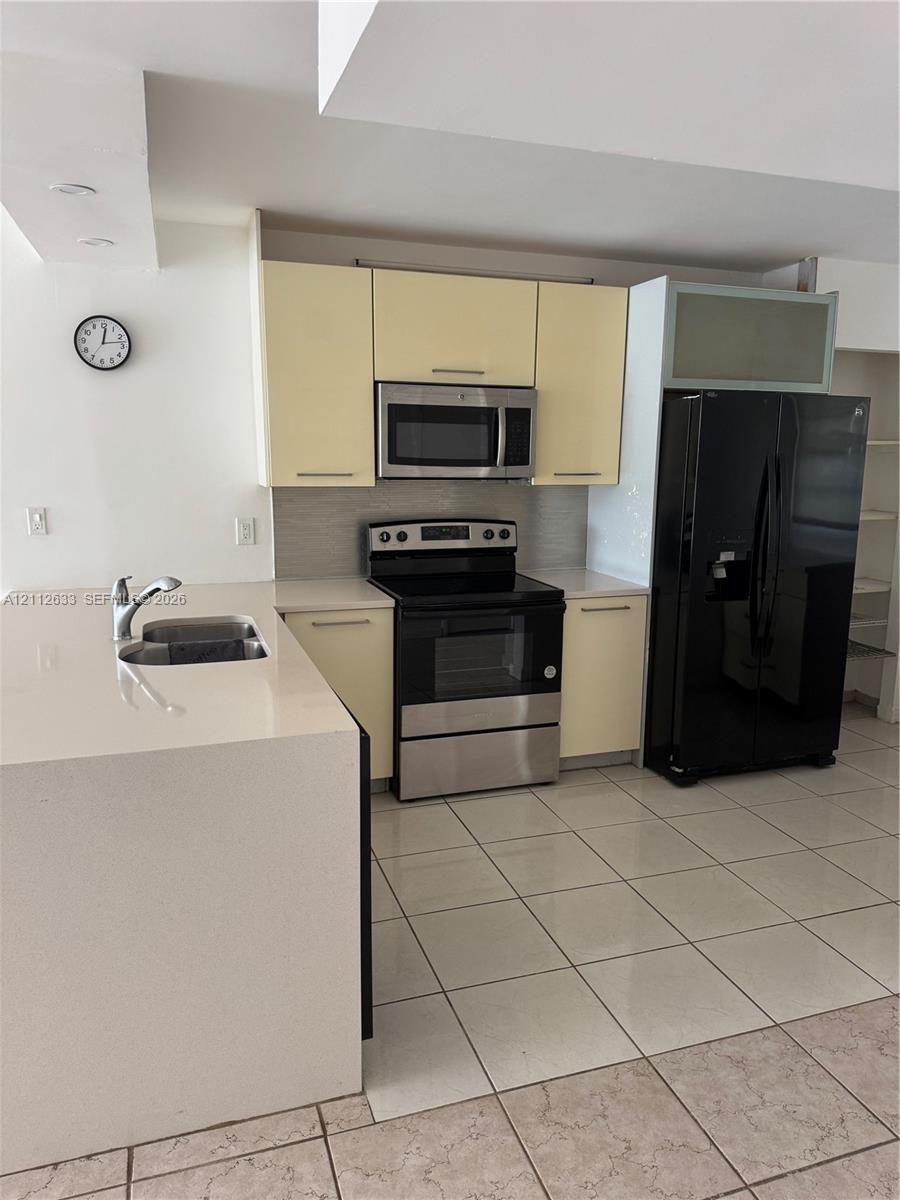 Kitchen featuring pale cabinetry, a stainless steel electric range, a microwave, a large-format refrigerator, and a solid-surface peninsula with an integrated sink
