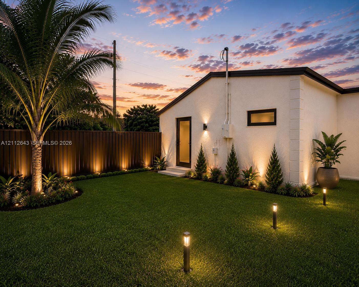 Landscaped backyard featuring a manicured lawn, established palm tree, structural exterior lighting, and private metal fencing Landscaped backyard featuring a manicured lawn, established palm tree, structural exterior lighting, and private metal fencing