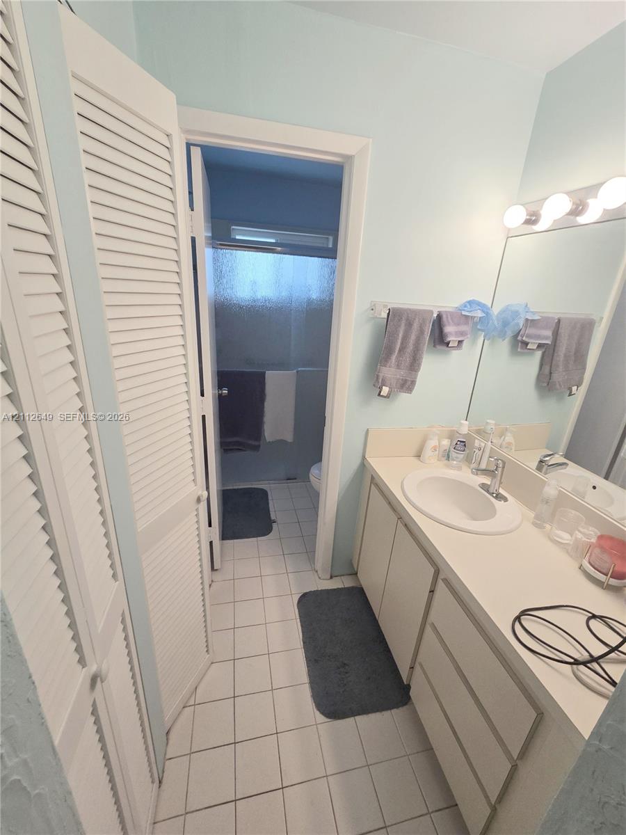 Bathroom featuring a single-sink vanity with white cabinetry, louvered closet doors, tile flooring, and a wall-mounted light fixture above a large mirror Bathroom featuring a single-sink vanity with white cabinetry, louvered closet doors, tile flooring, and a wall-mounted light fixture above a large mirror