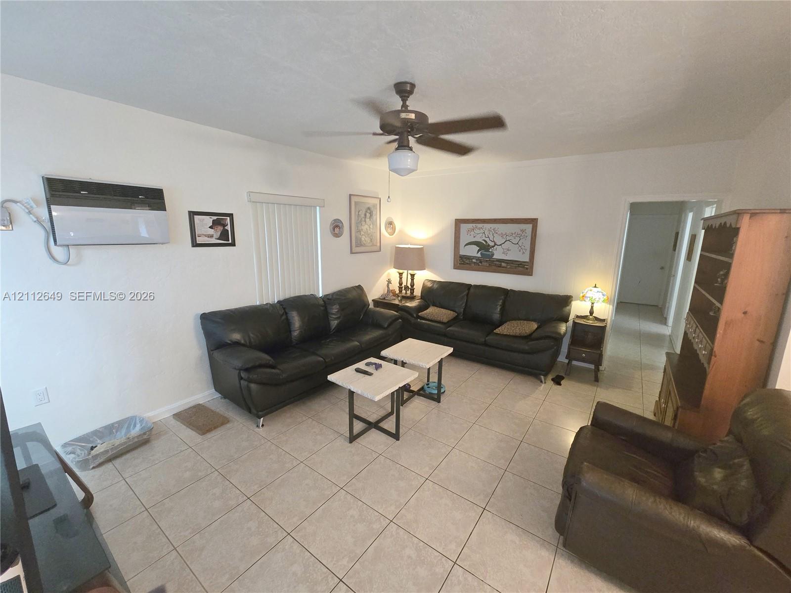 Room interior featuring tile flooring, ceiling fan, wall-mounted climate control unit, and vertical window blinds Room interior featuring tile flooring, ceiling fan, wall-mounted climate control unit, and vertical window blinds