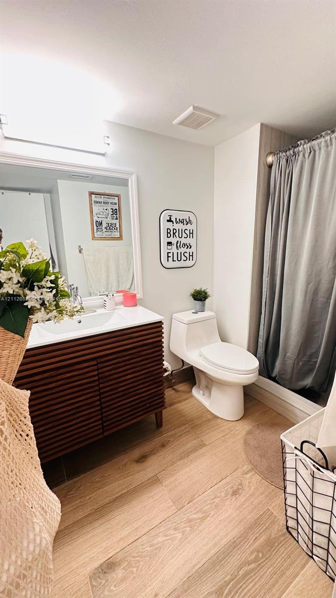 Bathroom featuring a wood-finish vanity with a white countertop and integrated sink, a rectangular framed mirror, a one-piece toilet, and wood-finish flooring