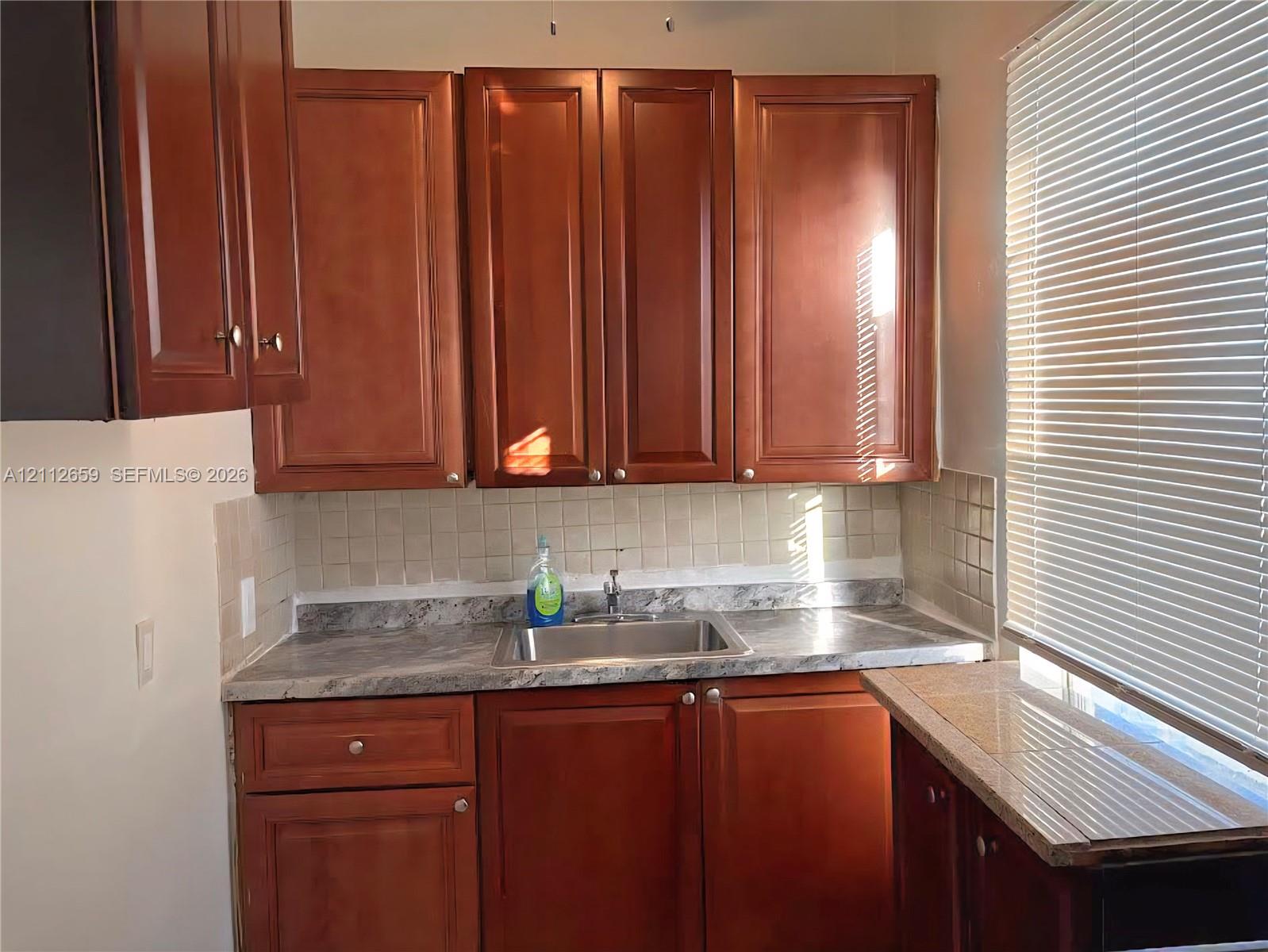 Kitchen area featuring wood-finish cabinetry, stone-patterned laminate countertops, square tile backsplash, and a drop-in stainless steel sink