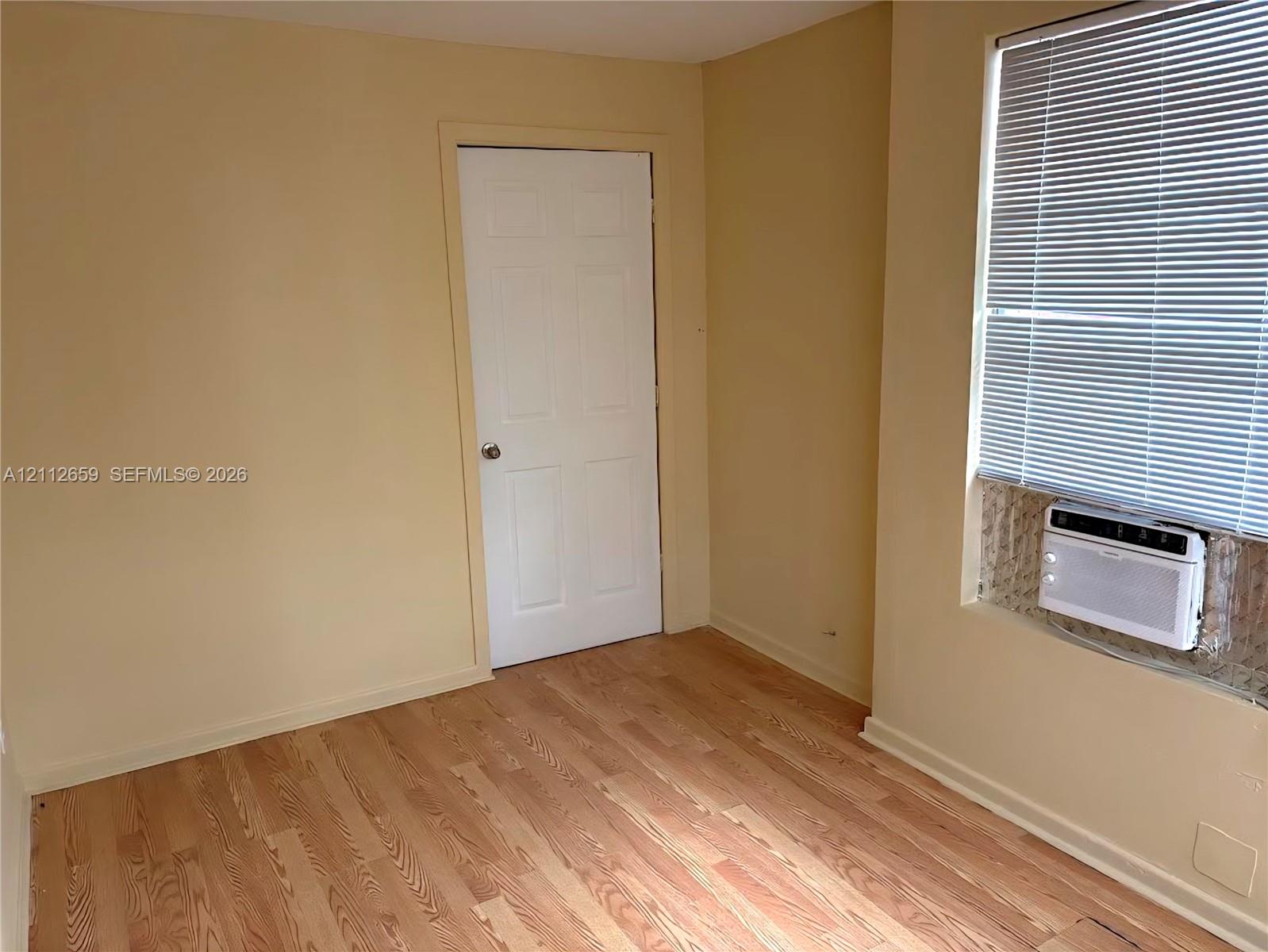 Room featuring wood-finish flooring, a six-panel door, and a wall-mounted air conditioning unit set within a window with horizontal blinds Room featuring wood-finish flooring, a six-panel door, and a wall-mounted air conditioning unit set within a window with horizontal blinds