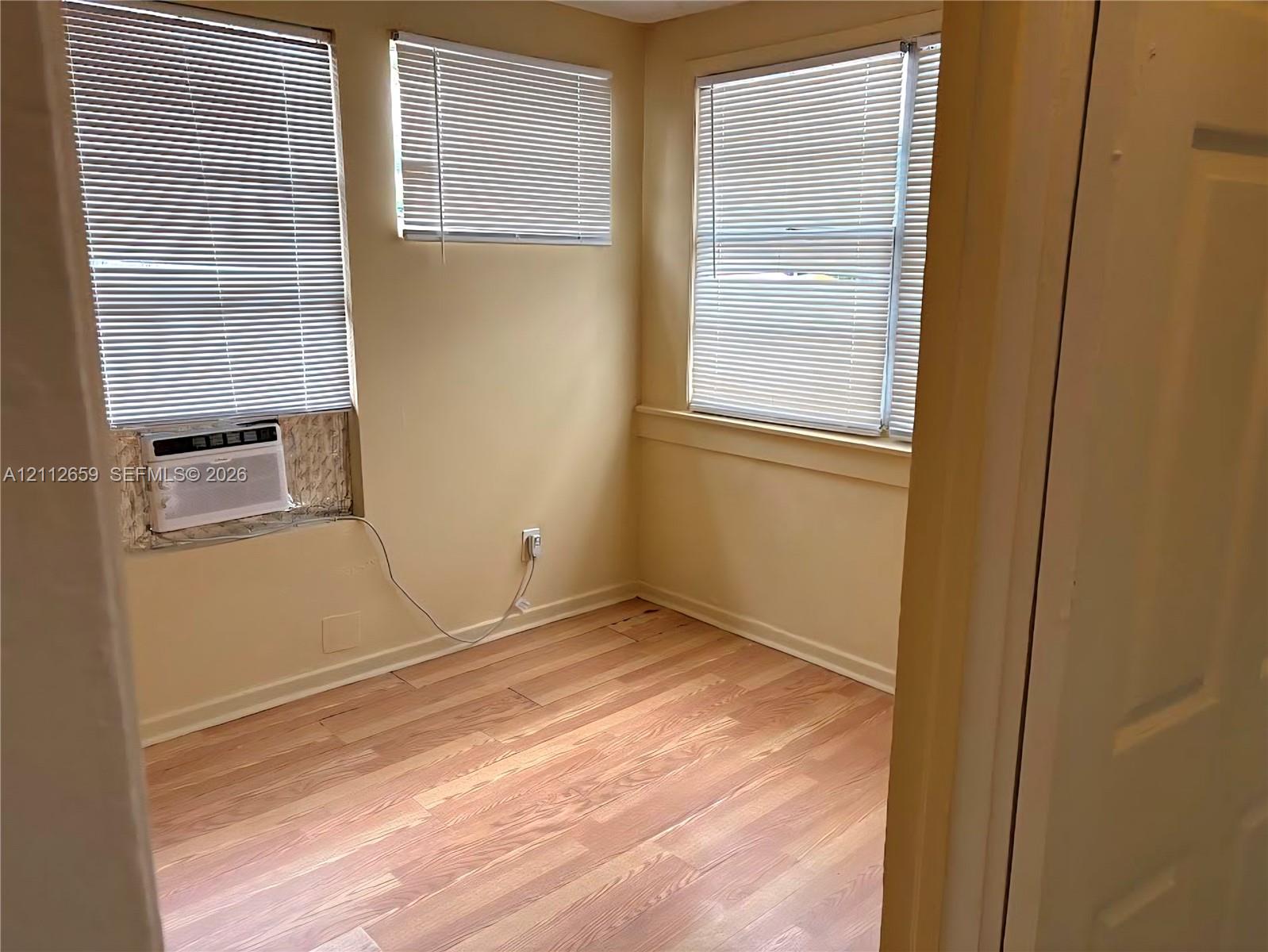 Room interior featuring wood-finish flooring, three windows with horizontal blinds, and a built-in wall-mounted air conditioning unit Room interior featuring wood-finish flooring, three windows with horizontal blinds, and a built-in wall-mounted air conditioning unit
