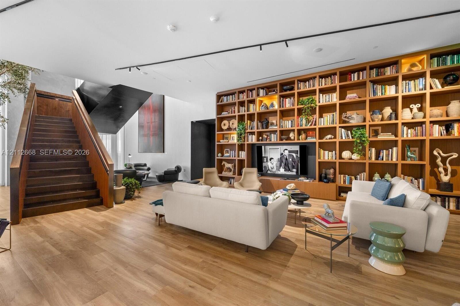 Expansive room featuring wood-finish flooring, floor-to-ceiling built-in wooden shelving, a floating-style staircase with wood treads, and recessed track lighting