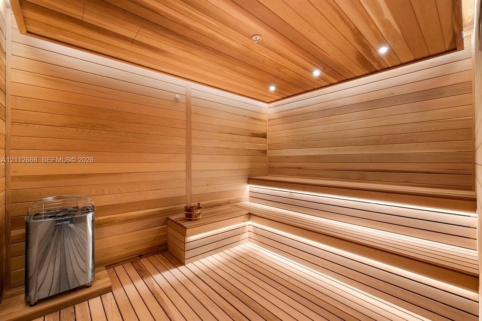 Sauna featuring light-toned wood wall and ceiling paneling, multi-level bench seating, integrated strip lighting, and a metal sauna heater
