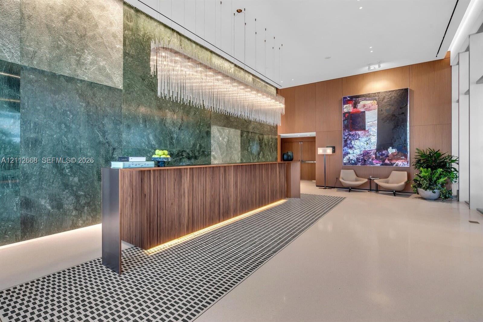 Grand lobby featuring a floor-to-ceiling polished stone accent wall, custom wood reception desk with integrated lighting, expansive linear crystal chandelier, wood-paneled walls, and geometric patterned inlay flooring