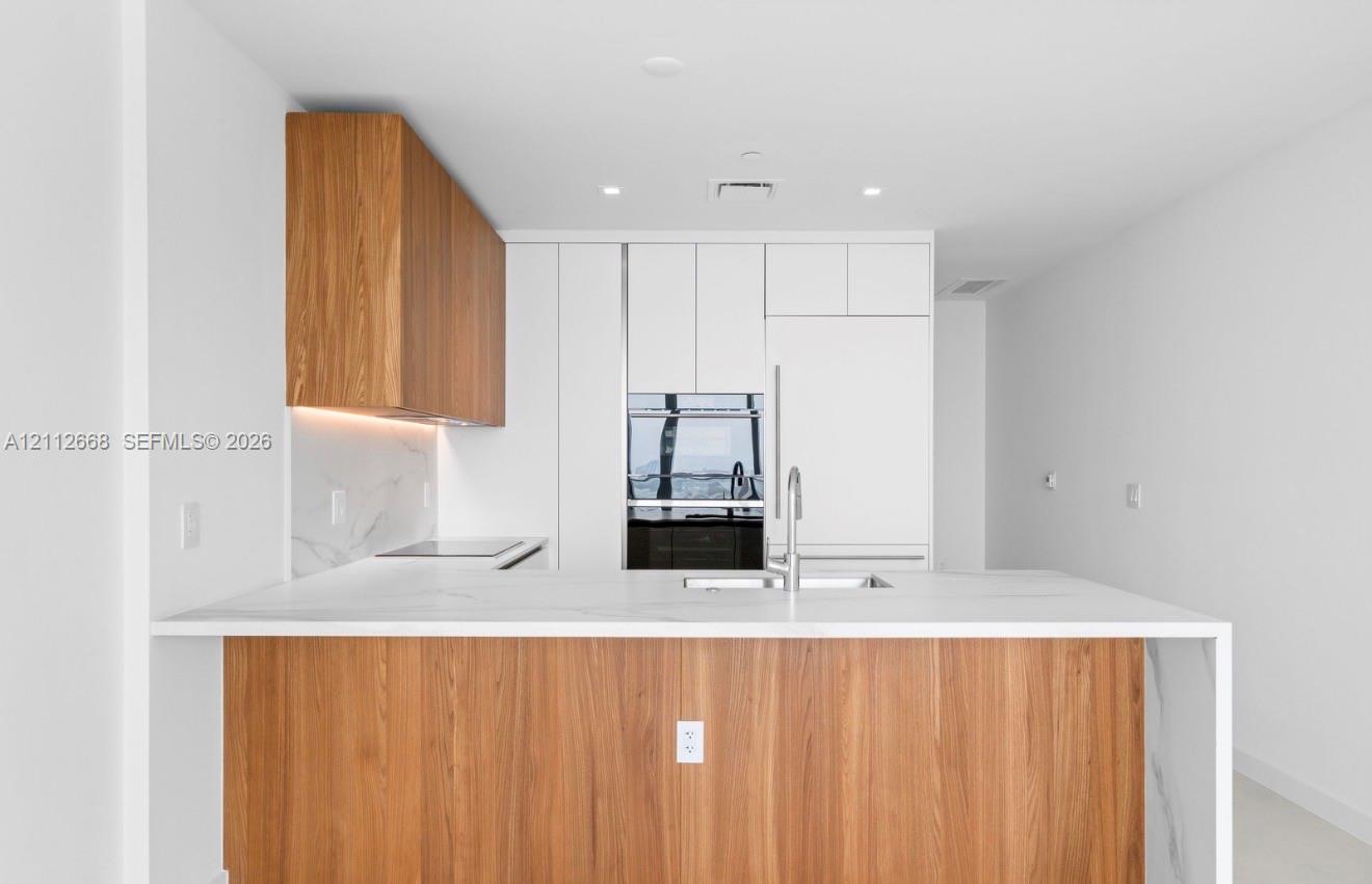 Modern kitchen featuring custom wood-finish cabinetry, white flat-panel cabinetry, waterfall-edge stone countertops, integrated appliances, and a stainless steel gooseneck faucet