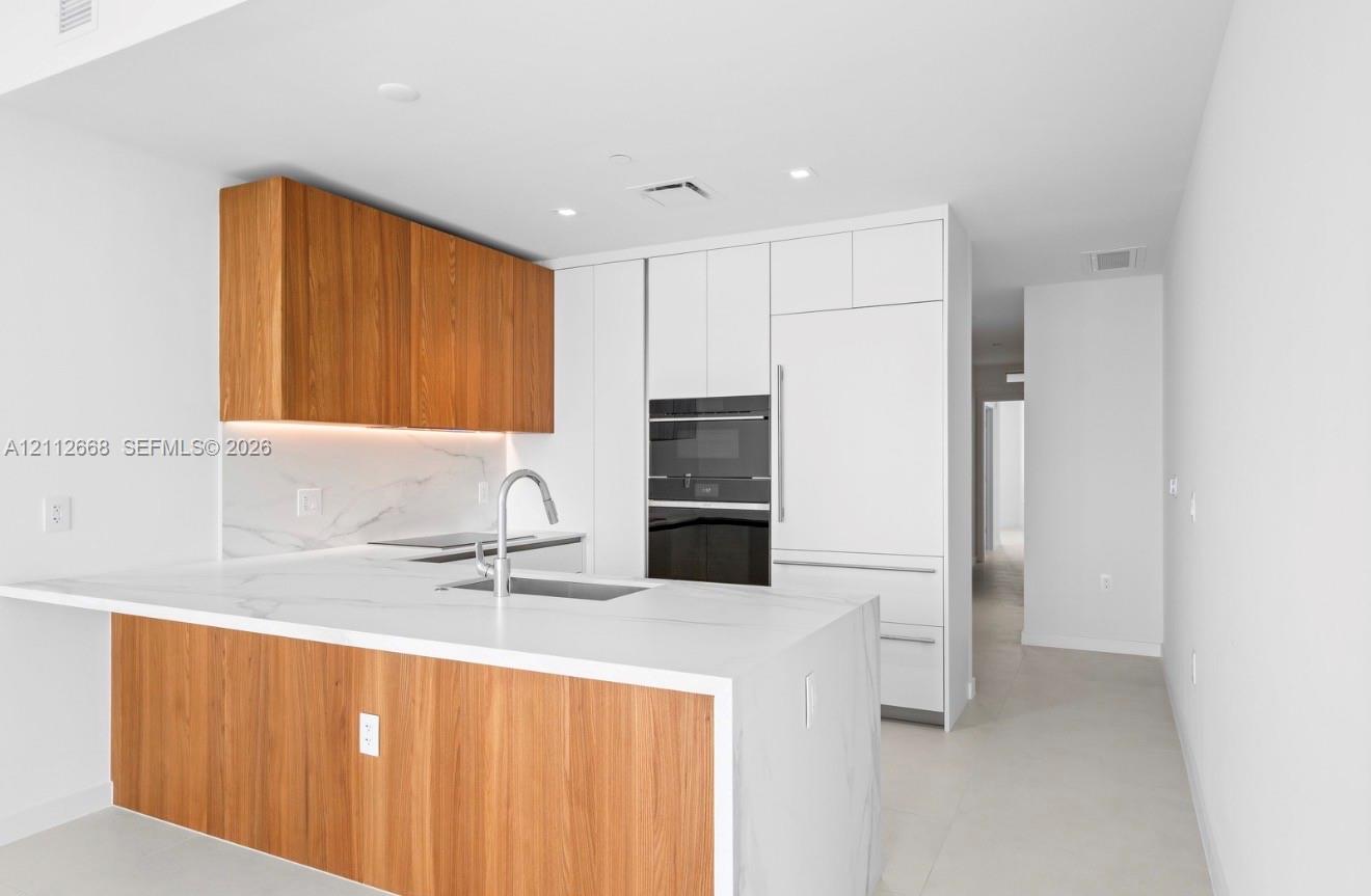 Kitchen featuring flat-panel wood-finish cabinetry, white solid surface countertops with integrated backsplash, built-in panel-ready refrigerator, stainless steel undermount sink with arched faucet, and stacked wall oven
