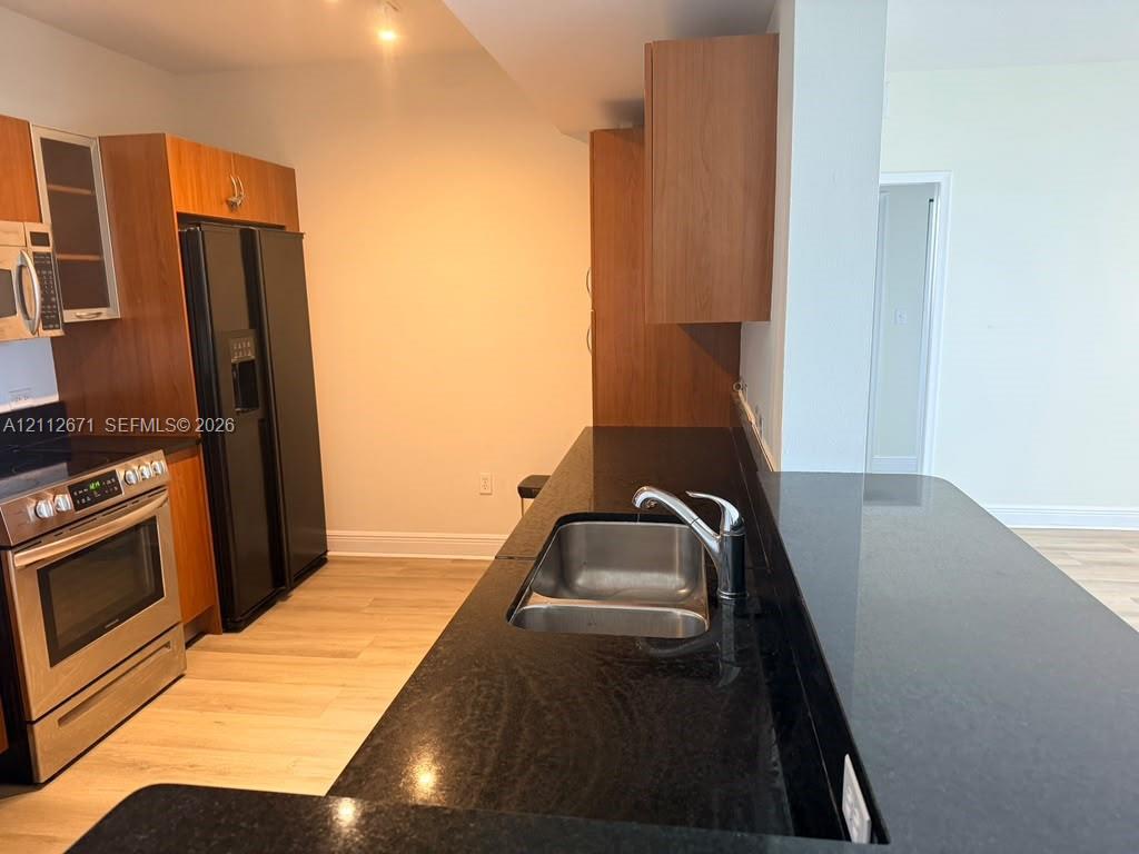 Kitchen featuring wood-finish cabinetry, dark stone countertops, stainless steel appliances, and dual-basin stainless steel sink with high-arc faucet Kitchen featuring wood-finish cabinetry, dark stone countertops, stainless steel appliances, and dual-basin stainless steel sink with high-arc faucet