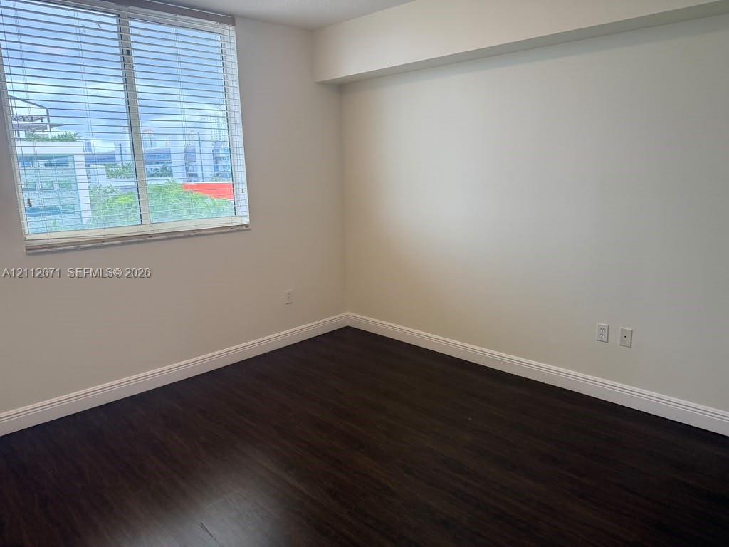 Neutral interior featuring dark wood-finish flooring, a large window with horizontal blinds, and smooth, light-toned walls Neutral interior featuring dark wood-finish flooring, a large window with horizontal blinds, and smooth, light-toned walls